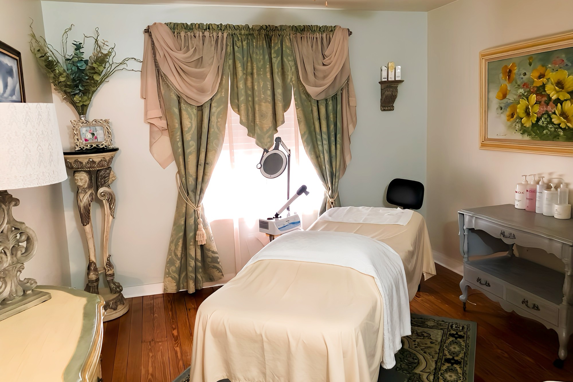 A cozy spa room with soft lighting, featuring a treatment bed covered with white linens in the center. Behind the bed, there are elegant green curtains with beige draping over a window. Floral decorations, a lamp, and beauty products are present in the room.